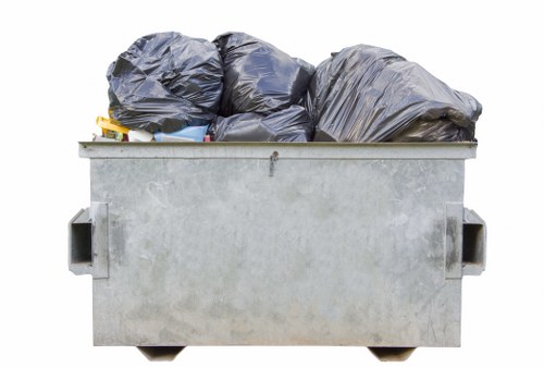 A large, rectangular metal dumpster with a flat, open top filled with multiple black plastic rubbish bags, some bulging and others tightly packed, all placed outdoors possibly on a driveway or near a garden area. The metal surface of the container shows a matte, slightly weathered finish with minor signs of use, and the black bags appear to contain mixed waste materials. The dumpster has sturdy, rectangular side handles for lifting or moving. In the background, there is no visible scenery or environment detail, emphasizing the focus on the waste collection. The image conveys the collection of garden or household waste typical of rubbish removal services in Hammersmith, with the placement suggesting preparedness for disposal or collection arranged by Garden Clearance Hammersmith. The scene emphasizes the practical aspect of rubbish disposal in an urban or suburban setting, with an emphasis on waste management and clearance solutions available locally.