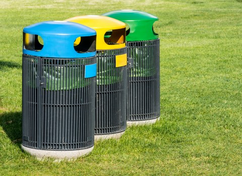 A row of three cylindrical outdoor rubbish bins placed on a grassy area, each with a black plastic body featuring vertical slats for ventilation and durability. The bins have smooth, rounded lids in bright yellow, blue, and green, with a large opening for waste disposal. The grassy ground beneath the bins is well-maintained with short, green turf, and the background shows more grass extending beyond the bins, indicating an open outdoor space or garden setting. These waste containers are typical for use in residential or public areas in Hammersmith, suitable for rubbish collection services provided by companies like Garden Clearance Hammersmith. The lighting suggests natural daylight, highlighting the vivid colors of the lids and the texture of the grass, creating a clean and orderly scene that visually emphasizes waste management in a local environment.