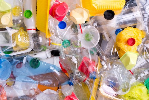 A close-up view of a large pile of discarded plastic bottles, caps, and containers, some crumpled or crushed, with various colors including clear, yellow, red, green, blue, and black. The bottles are made of translucent and opaque plastics with different textures, including smooth surfaces and ridged sections. The background shows a mixture of crushed and intact bottles, some with labels partially visible, resting on a flat surface likely outdoors or in a waste collection area. The arrangement appears random, with some bottles overlapping and others leaning against each other, creating a chaotic but identifiable collection of plastic waste. This image presents a visual of domestic or commercial plastic rubbish, typical of waste collected for recycling or disposal by rubbish removal services, such as those offered by Garden Clearance Hammersmith in the Hammersmith area, which operates within the local postcode district.
