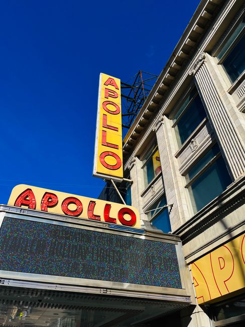 A tall, vertical yellow sign with red letters reading 'APOLLO' is mounted on the side of a historic building with decorative stonework and large windows. Below the sign, a marquee with the word 'APOLL