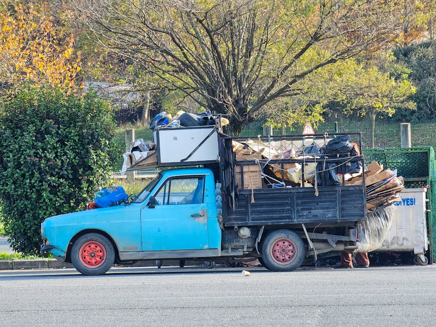 A street scene in an urban area shows a waste collection vehicle positioned alongside a row of older buildings with mixed facade finishes, some showing signs of weathering or partial renovations. The 
