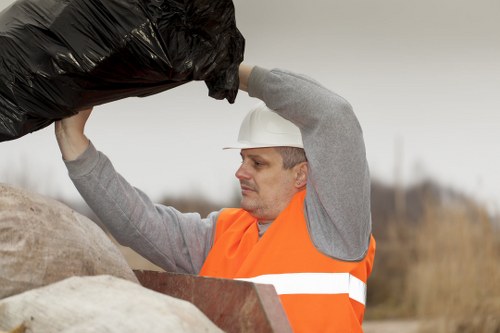 A male waste collection worker wearing a white safety helmet and an orange high-visibility vest is lifting a black rubbish bag over his shoulder in an outdoor rural setting. The background features a cloudy sky with bare trees and open land, suggesting a countryside or garden environment. The worker is standing next to a large pile of mixed waste materials, including brown and beige sacks and some debris, likely awaiting collection or disposal. His focused expression indicates careful handling of refuse. This scene relates to rubbish removal services offered by Garden Clearance Hammersmith, with the image emphasizing manual waste collection in an open, natural area typical of garden and outdoor clearance work in the Hammersmith area or nearby postcodes. The textures of the rubbish bag appear crinkled and durable, contrasting with the smooth fabric of the worker’s safety gear. The overall environment is practical and professional, reflecting waste management activities relevant to local garden clearance and rubbish removal services.
